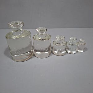 May include: A set of five clear glass scale weights arranged in descending order of size. The weights are labeled 1kg, 1/2kg, 200g, 100g, and 50g. Each weight features a cylindrical body with a rounded neck and a flat top knob for handling.