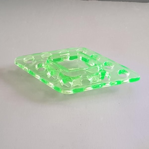 May include: A square, neon green glass ashtray with a central square and circular indentations. The ashtray has a diamond shape with rounded edges and is made of transparent glass.