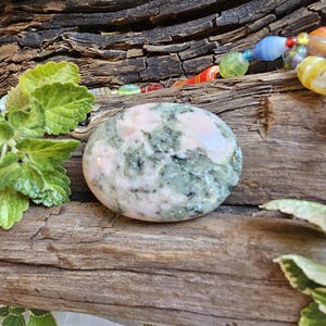 May include: A polished, oval-shaped stone featuring a mottled pattern of soft pink and sage green mineral inclusions. The smooth, rounded surface displays a natural variation of colors and textures, resting alongside green leaves and colorful beads.