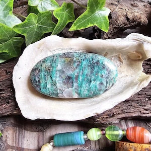 May include: An oval-shaped, polished gemstone with a mix of teal and green hues, resting in a white, shell-like structure. The stone's surface displays a unique pattern of lines and textures. Green ivy leaves and a colorful beaded necklace are also visible.