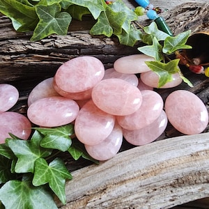 May include: A collection of smooth, polished, oval-shaped rose quartz stones with a translucent pale pink hue. The stones are arranged in a cluster, showcasing their glossy, reflective surfaces and natural variations in color and internal texture.