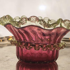 Può includere: Una ciotola vintage in vetro color rubino con bordo a balze. La ciotola ha una forma svasata con un bordo decorativo smerlato in una tonalità verde più chiara. Anche la base della ciotola presenta un design decorativo smerlato. Il vetro ha una finitura lucida e riflettente.