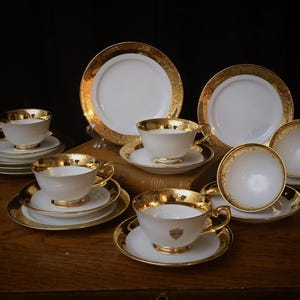 Puede incluir: Juego de té blanco y dorado. El conjunto incluye tazas, platillos y platos con un diseño de filigrana dorada alrededor de los bordes. Las tazas tienen asas doradas y un escudo. El juego está dispuesto sobre una superficie de madera.