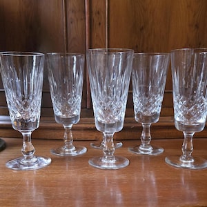 May include: Five clear crystal wine glasses with a diamond pattern on the lower half and vertical lines on the upper half. The glasses are displayed on a wooden surface. The background features a dark wooden cabinet.