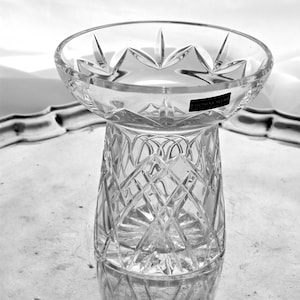 Puede incluir: Un jarrón de cristal tallado transparente con un borde ancho en forma de estrella y una base con diseño de diamantes. El jarrón se encuentra sobre una superficie reflectante, mostrando su intrincado diseño y claridad. La etiqueta dice "THOMAS WEBB".