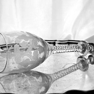 May include: A clear, etched glass with a floral design, lying on a silver tray. The stem has a twisted design, and the glass is reflected in the tray's surface. The background is a white, draped fabric.
