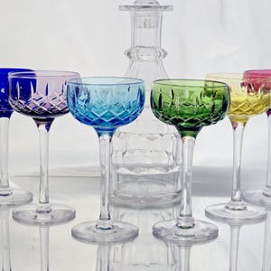 Può includere: Un set di sei coppe in cristallo multicolore blu, viola, azzurro, verde, giallo e magenta, ciascuna con un motivo a taglio diamante sulla coppa e uno stelo trasparente, disposte davanti a un decanter in vetro trasparente con motivo abbinato.