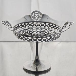 May include: A silver-colored, ornate serving dish with a pedestal base. The dish features a pierced, openwork design around the bowl and decorative flourishes. The base is round and the stem is slender.