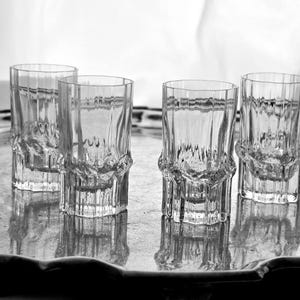 May include: Four clear shot glasses with a textured design, placed on a silver tray. The glasses have a distinctive base and a ribbed pattern, reflecting light and creating shadows. The image is in monochrome.