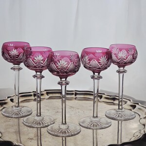 Può includere: Cinque calici da vino con coppa rosa intenso e stelo trasparente. I calici presentano un motivo in cristallo intagliato e sono disposti su un vassoio d'argento. Lo sfondo è bianco.