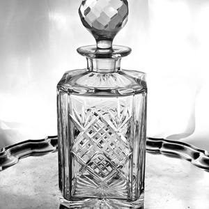 May include: A clear, cut-glass decanter with a geometric design and a faceted stopper. The rectangular decanter features a diamond pattern. It is displayed on a silver tray, reflecting light. The decanter is likely for spirits or wine.