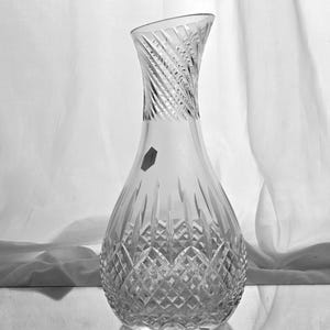 May include: A clear glass decanter featuring a wide, rounded base with a diamond-cut pattern and a slender, tapered neck with diagonal etched lines. The vessel has a slanted rim and a small, dark hexagonal sticker on its upper body.