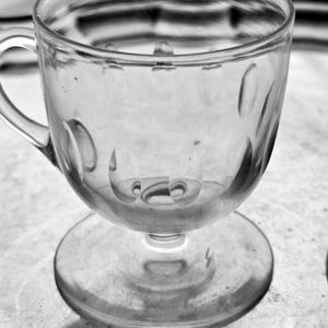 May include: A clear, stemmed glass teacup with a handle. The cup has a rounded bowl with decorative oval indentations. The base is a flat, circular foot. The glass is transparent, allowing light to pass through.