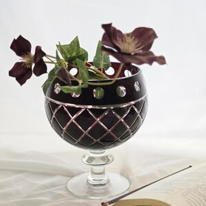 May include: A dark red crystal goblet with a diamond pattern and a clear stem, holding dark purple flowers and green leaves. The goblet has a silver rim and is set on a white surface, next to an open book.