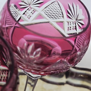 May include: Close-up of a pink crystal wine glass with intricate white cut designs. The glass has a clear stem and a wide bowl, reflecting light and the surrounding environment. The cut patterns include geometric shapes and floral motifs.