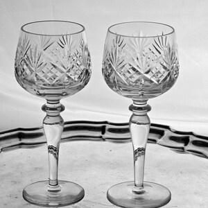 May include: Two clear crystal wine glasses with detailed cut glass designs. The glasses feature a rounded bowl, a delicate stem, and a circular foot. They are presented on a reflective silver tray, offering a classic and elegant appearance.