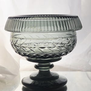 May include: A gray glass pedestal bowl with a decorative rim and diamond-cut pattern. The bowl sits on a tiered base. The glass has a reflective surface.