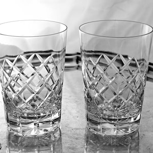 May include: Two clear, cut-glass tumblers with a diamond pattern sit on a reflective surface. The glasses are cylindrical with a thick base and a smooth rim. The image is in black and white.