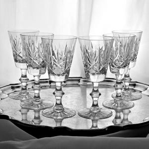 Op de afbeelding: Zes kristallen wijnglazen met stervormige patronen staan op een zilveren serveerschaal. De glazen hebben een voet en een uitlopende kelk. De schaal heeft een decoratieve, geschulpte rand die het licht reflecteert. De glazen zijn ongeveer 15 cm hoog.