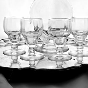 May include: A set of clear glass stemware, comprising four small goblets and a decanter, presented on a silver-toned tray. The goblets feature a detailed base and rounded bowl. The glassware reflects light, creating a classic, elegant aesthetic. The tray is silver-toned.