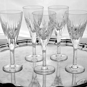 May include: Five clear crystal wine glasses with etched vertical lines, arranged on a silver-colored, ornate tray. The glasses have a slender stem and a flared bowl. The tray has a decorative, scalloped edge.