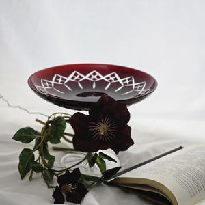 Può includere: Una ciotola di vetro rosso rubino con un motivo geometrico bianco sul bordo. La ciotola è sollevata su un supporto di vetro trasparente. Fiori bordeaux scuro e un libro sono disposti nelle vicinanze su un panno bianco.