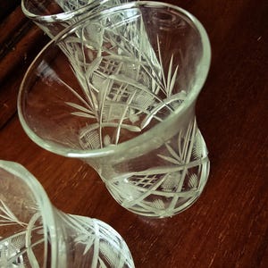 Op de afbeelding: Close-up van drie heldere, geëtste glazen bekers. De glazen hebben een uitlopende vorm en ingewikkelde geometrische en bloemmotieven. De glazen staan op een donkerbruin houten oppervlak.
