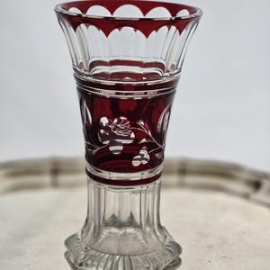 Può includere: Un vaso di vetro trasparente con bordo e dettagli rosso rubino. Il vaso presenta un bordo smerlato e disegni floreali incisi. La base è un design trasparente e sfaccettato. Il vaso è esposto su un vassoio d'argento.