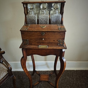 May include: An antique wooden vanity table with three glass bottles. The bottles have crystal stoppers and are displayed in a mirrored compartment. The table has a small drawer and ornate, curved legs. The wood has a warm, brown tone.