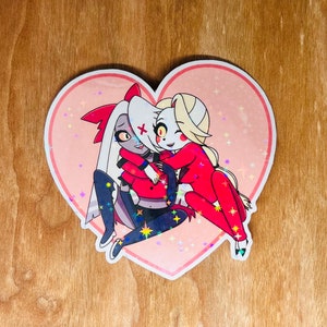 Puede incluir: Una pegatina en forma de corazón con fondo rosa y purpurina. Dos personajes de dibujos animados se abrazan dentro del corazón. El personaje de la izquierda lleva un vestido rojo y pantalones negros. El personaje de la derecha lleva un traje rojo y tiene el pelo rubio.