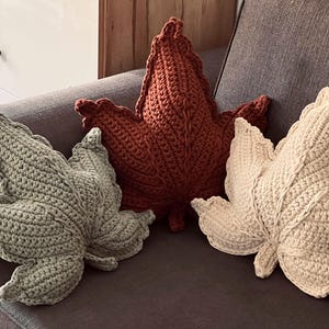 Puede incluir: Tres cojines con forma de hoja elaborados con un punto de ganchillo texturizado. Los cojines son de color verde salvia, naranja quemado y crema. Cada cojín presenta un detalle de vena central y un borde festoneado, imitando una hoja de arce.