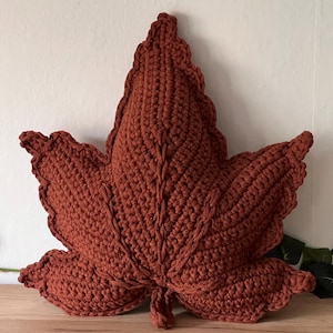 May include: A rust-colored, leaf-shaped throw pillow crafted with a textured crochet stitch. The pillow features a prominent central vein and detailed, scalloped edges that mimic the natural structure of a maple leaf in a solid, deep orange-brown tone.