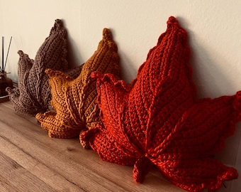 May include: Three leaf-shaped crochet pillows in autumn colors. The front pillow is bright orange, the middle pillow is mustard yellow, and the back pillow is dark brown. Each pillow features a textured, ribbed stitch pattern mimicking leaf veins.