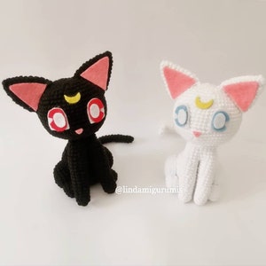May include: Two crocheted cat dolls, one black and one white, each featuring large pointed ears with pink felt inserts, a yellow crescent moon on the forehead, and large eyes. The text @lindamigurumis is visible between the two figures.