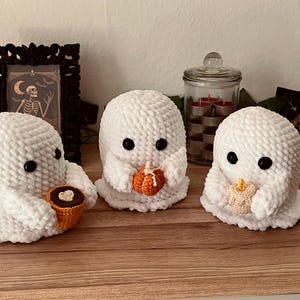 Könnte beinhalten: Drei gehäkelte Geisterfiguren aus weichem Garn in Weiß. Jeder Geist hat schwarze Knopfaugen. Der linke Geist hält eine braune Tasse mit Herz, der mittlere Geist einen kleinen orangefarbenen Kürbis und der rechte Geist eine beige Kerze.
