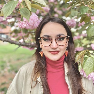 Op de afbeelding: Een vrouw met lang bruin haar draagt een rode trui met een hoge hals en een beige jas. Ze draagt een zwarte cat-eye bril en pizza-plak oorbellen. Ze staat voor een roze kersenbloesemboom.