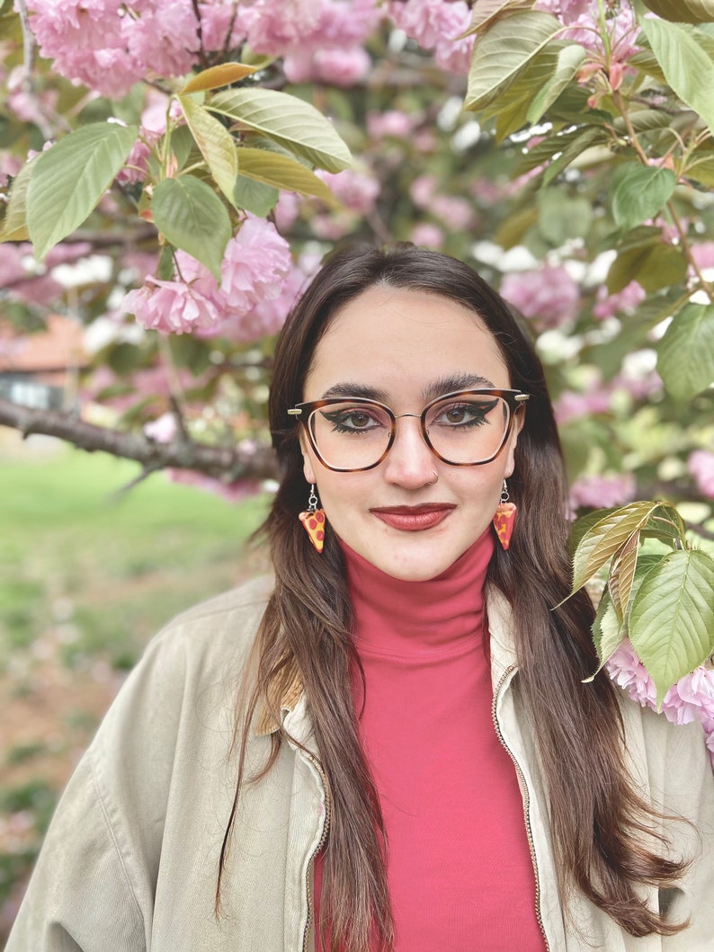 Op de afbeelding: Een vrouw met lang bruin haar draagt een rode trui met een hoge hals en een beige jas. Ze draagt een zwarte cat-eye bril en pizza-plak oorbellen. Ze staat voor een roze kersenbloesemboom.