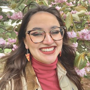 Op de afbeelding: Een vrouw met lang bruin haar lacht en draagt een rode trui met een hoge hals, een beige jas en een bril. Ze draagt ​​pizza-oorstekers. De achtergrond is een wazig beeld van roze bloemen.