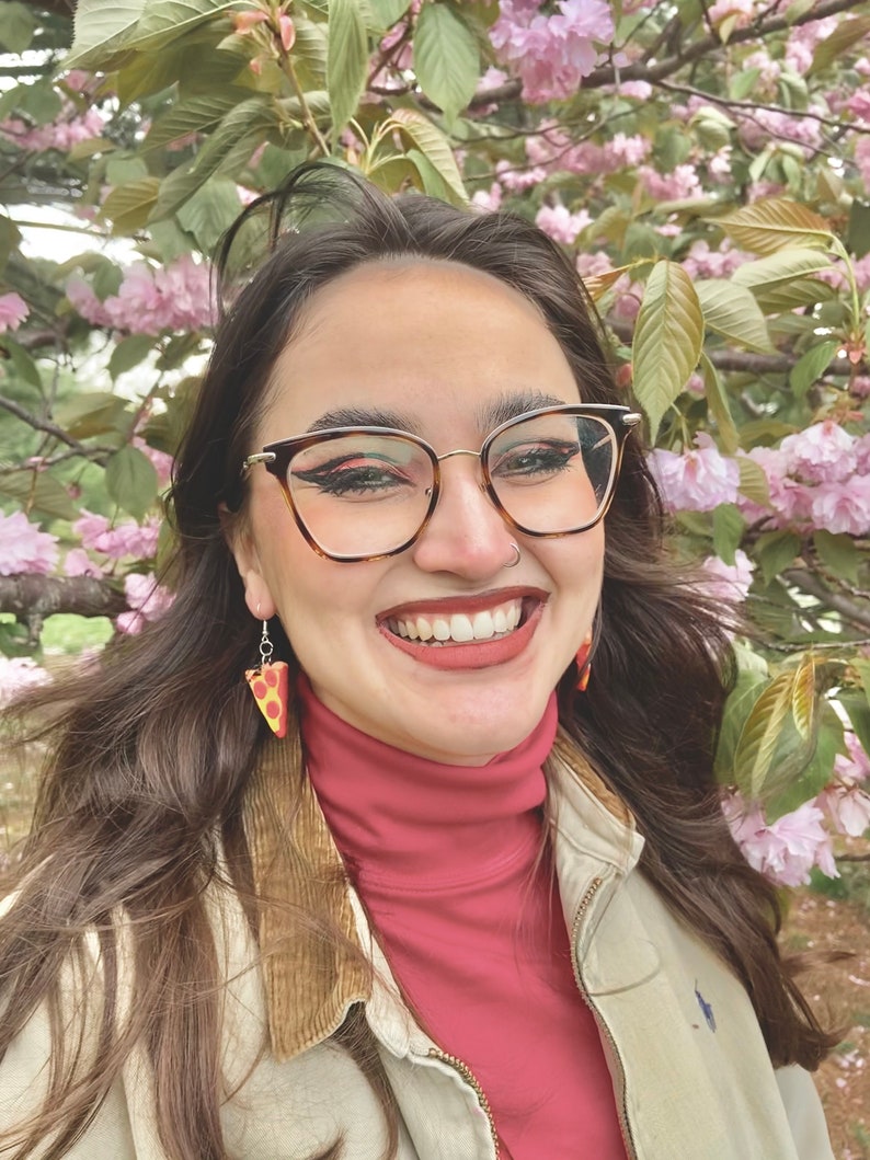 Op de afbeelding: Een vrouw met lang bruin haar lacht en draagt een rode trui met een hoge hals, een beige jas en een bril. Ze draagt ​​pizza-oorstekers. De achtergrond is een wazig beeld van roze bloemen.