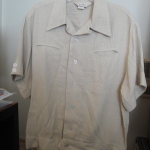 May include: A beige short-sleeved button-down shirt with a textured fabric. The shirt has a collar and a pocket on each side. The shirt has a label that says "The Shirt Shop".