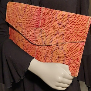 May include: A rectangular clutch with a reptile-like pattern in shades of red and orange. The clutch has a flap closure and a circular accent. The background is a black garment.