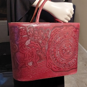 May include: Red leather tote bag with embossed design. Features a detailed image of a person on horseback, cacti, and a circular calendar. The bag has two handles and a rectangular shape.