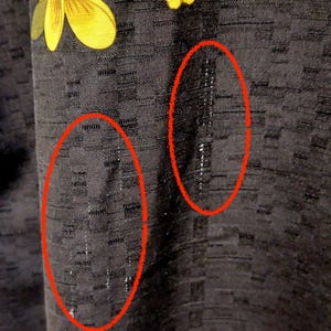 May include: Close-up of a black fabric with a textured, grid-like pattern. The fabric has a slight sheen and appears to be slightly wrinkled in areas. A yellow floral design is visible in the upper left corner.