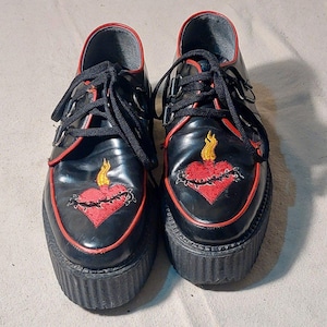 May include: Pair of black platform shoes with red trim and laces. Each shoe features an embroidered red heart with a yellow flame and thorny vines. The shoes have a thick, black sole.