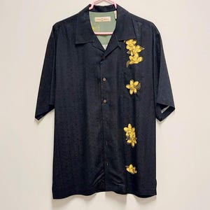 May include: Black button-down shirt with short sleeves and a floral pattern. The shirt features yellow flower designs on the front. The shirt is hanging on a pink hanger.