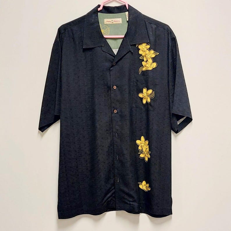 May include: Black button-down shirt with short sleeves and a floral pattern. The shirt features yellow flower designs on the front. The shirt is hanging on a pink hanger.