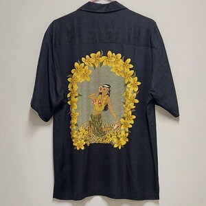May include: Dark blue short-sleeved shirt featuring a vibrant illustration of a woman in a floral garland, surrounded by yellow flowers. The artwork is centered on the back of the shirt, creating a tropical theme. The shirt has a classic collar and button-up design.