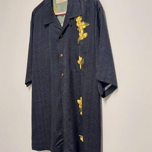 May include: A dark navy blue button-down shirt with short sleeves. The shirt features a subtle textured pattern and is adorned with yellow floral embroidery on the front. The shirt is hanging on a pink hanger.