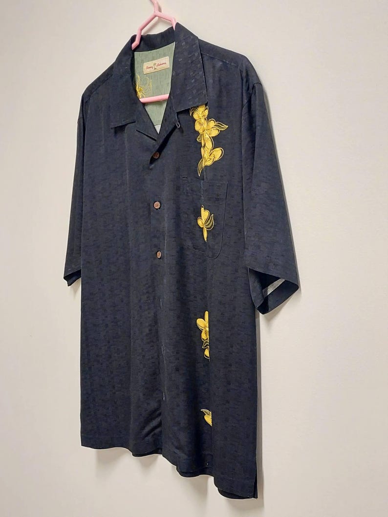 May include: A dark navy blue button-down shirt with short sleeves. The shirt features a subtle textured pattern and is adorned with yellow floral embroidery on the front. The shirt is hanging on a pink hanger.