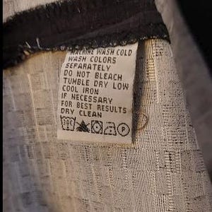 May include: Close-up of a clothing tag with care instructions. The tag is white with black text, including "MACHINE WASH COLD," "TUMBLE DRY LOW," and "DRY CLEAN." The fabric has a textured, woven pattern in shades of gray and white.